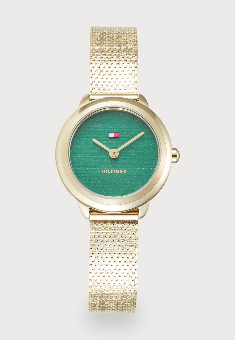 Gouden horloge met een gestructureerde metalen meshband, met een groene wijzerplaat met subtiele glans, twee wijzers en het "Hilfiger"-logo.
