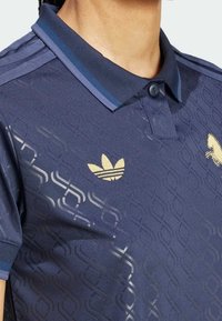 Tummansininen polo-paita, jossa on tekstuurinen geometrinen kuvio, kultainen Adidas-logo vasemmassa rinnassa sekä kaksivärinen kaulus, jossa on sinisiä yksityiskohtia.