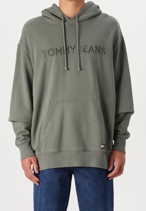Person wearing olive green hoodie with "TOMMY JEANS" embroidered on chest and small logo tag near kangaroo pocket, paired with blue jeans.