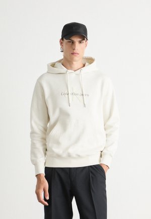 Sudadera crema con cordones y bolsillo delantero, con el logotipo bordado de "Calvin Klein Jeans" en color claro. Una gorra negra completa el look.