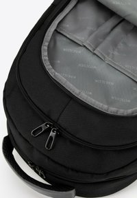 Un sac à dos noir avec un extérieur texturé, des zips doubles et une doublure intérieure grise présentant un motif de logo répété. Design compact et arrondi.
