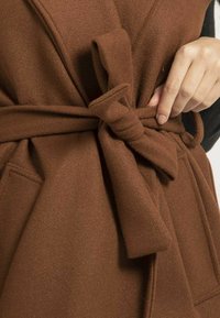 Enveloppe en polaire marron, douce, avec une taille nouée, présentant une texture lisse et des poches latérales. Le nœud est positionné à l'avant.