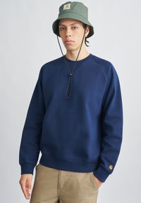 Μπλε ναυτικό φούτερ με ριμπ μανσέτες και τελείωμα, με λογότυπο στο στήθος. Φοριέται με πράσινο καπέλο τύπου κάπα και χακί παντελόνια.