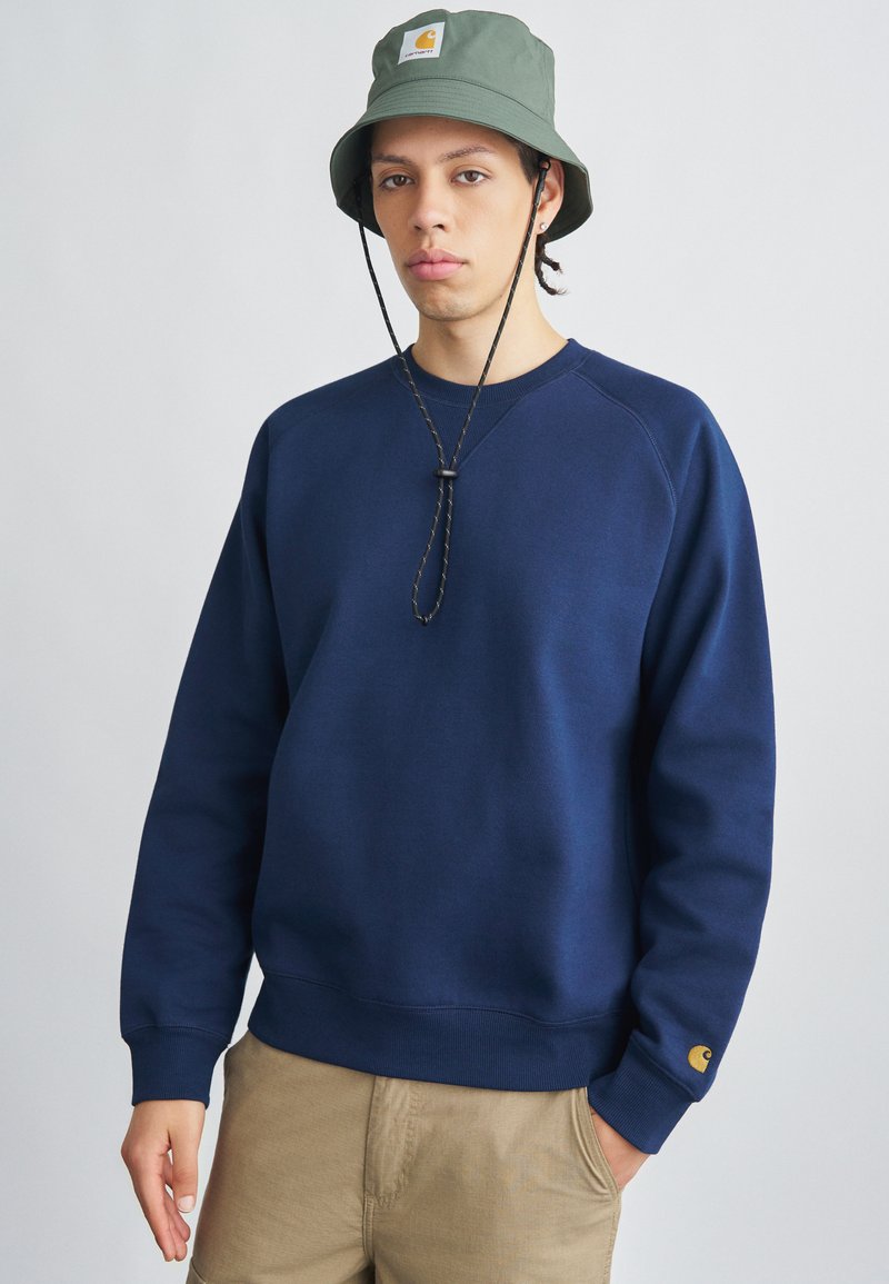 Marineblå sweatshirt med ribbet manchetter og kant, med et logo-patch på brystet. Båret med en grøn spandhat og khakibukser.