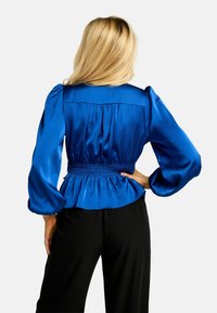 Saténová modrá blúzka s nafúknutými rukávmi, stiahnutá v páse a s volánovým lemom, zobrazená zozadu, nosená s čiernymi nohavicami.