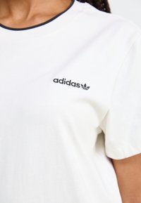 Valkoinen puuvillainen t-paita, jossa on pyöreä kaula-aukko ja tummansininen reunus, vasemmassa rinnassa pieni musta "adidas"-logo.