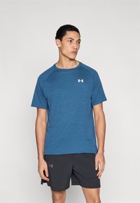 Μπλε αθλητική μπλούζα με κοντά μανίκια και στρογγυλή λαιμόκοψη. Διαθέτει διακριτή υφή και μικρό λευκό λογότυπο στο στήθος. Συνδυάζεται με μαύρο σορτς.