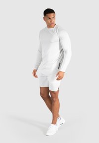 Camiseta deportiva de manga larga blanca con tejido texturizado y un logo lateral, combinada con pantalones cortos blancos y zapatillas blancas, sobre un fondo gris.