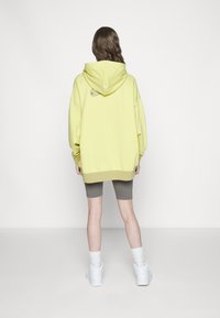 Felpa con cappuccio di colore giallo chiaro, vestibilità oversized, con un piccolo logo sul retro; abbinata a pantaloni corti grigi e sneakers bianche.