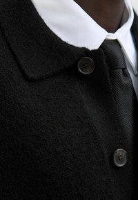 Melna teksturēta mētelis ar lieliem pogām, uzvilkts virs baltas kreklu ar apkakli un rakstainas melnas kaklasaites, tuvplāns uz augšējo krūšu un apkakles zonu.