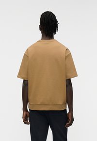 Καφέ βαμβακερό φανελάκι με κοντά μανίκια, στρογγυλή λαιμόκοψη και άνετη εφαρμογή, με ριμπ λεπτομέρειες στο κάτω μέρος. Όψη από πίσω.