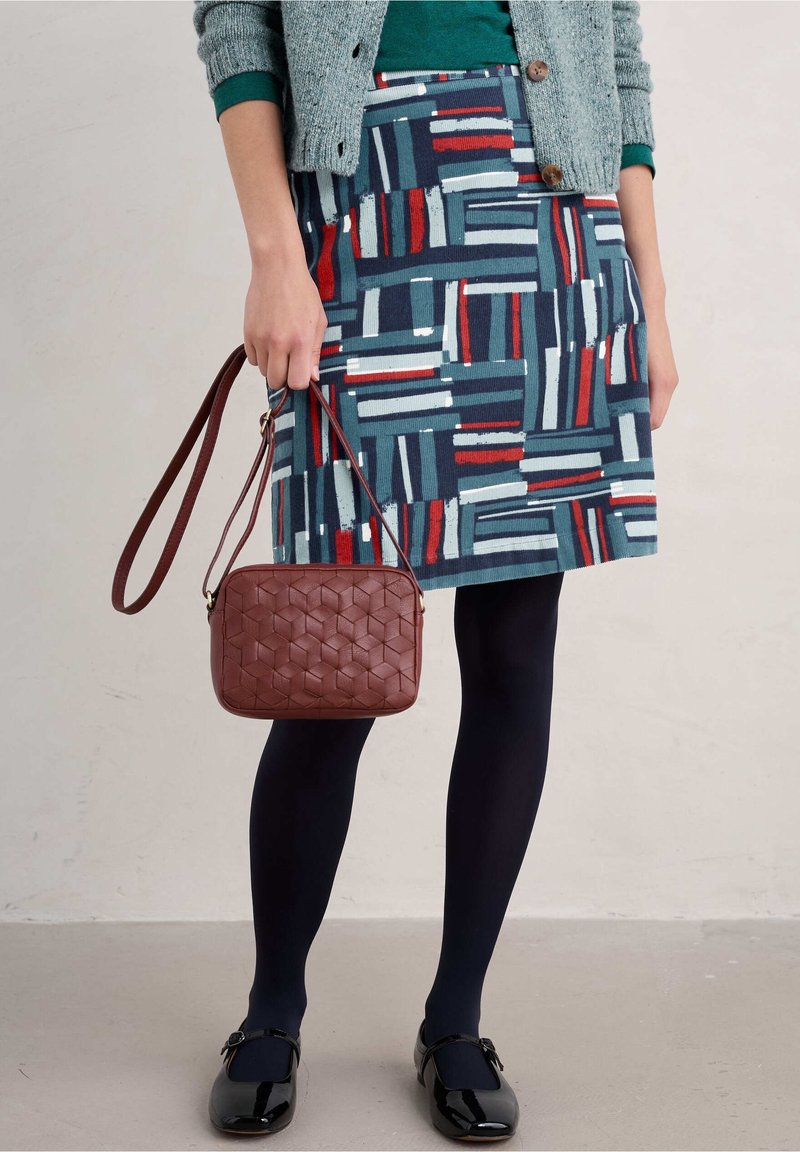 Patterned skirt in blue, red, and teal, paired with a brown woven bag, black shoes, and navy tights, layered under a green top and gray cardigan.