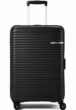 Schwarzer Hartschalenkoffer mit horizontalen Rippen, ausziehbarem Griff, vier Rollen und dem „American Tourister“-Logo auf der Vorderseite.