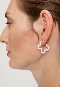 Weiße, abstrakt geformte Ohrringe mit einem fließenden Konturdesign, die eine glänzende Oberfläche und leichtes Material aufweisen, getragen am Ohr eines Modells.