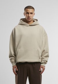 Haut en hoodie beige avec une grande poche avant, des manches larges et une capuche à cordon. Fabriqué dans un matériau doux, il a une coupe décontractée et une texture lisse.