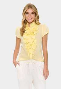 Blusa gialla con maniche corte a sbuffo, caratterizzata da un davanti arricciato e chiusura con bottoni. Tessuto testurizzato con un aspetto morbido e leggero.