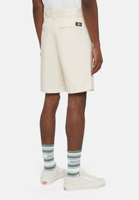 Pantaloni corti beige realizzati in un tessuto resistente, con una tasca posteriore e un'etichetta con il logo. Calzini rigati grigi e verdi e sneakers bianche completano il look.