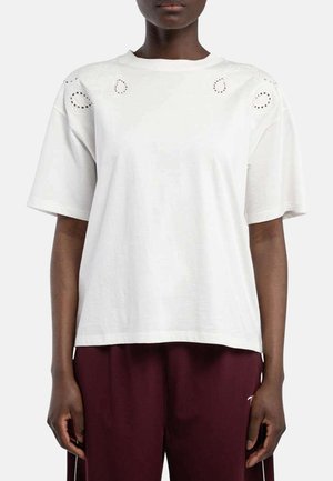 Une personne porte une chemise blanche ample à manches courtes avec des détails ajourés brodés près des épaules et un pantalon bordeaux foncé, debout devant un fond blanc.