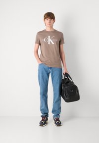 Rudas apvalios kaklo marškinėliai su balta "Calvin Klein Jeans" logotipu, derinami su šviesiai mėlynomis džinsais ir juoda sportine krepšiu. Batai su daugiaspalviu dizainu.