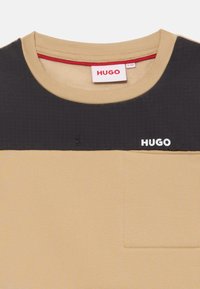 Camisa de manga corta beige y negra con cuello redondo, pequeño bolsillo en el pecho y logo blanco "HUGO" en el panel frontal superior negro.