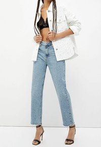 Chaqueta de denim blanca con un patrón moteado, sujetador de encaje negro, jeans de cintura alta azul claros y tacones altos negros con tiras.