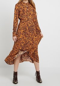 Abito a maniche lunghe di colore arancione con stampa floreale nera, caratterizzato da una chiusura con bottoni sul davanti, vita attillata e orlo asimmetrico. Indossato con stivaletti neri.
