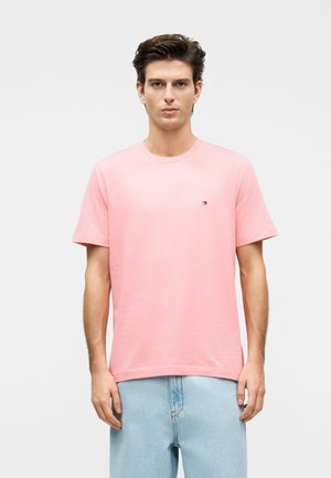 Mann mit braunen Haaren, der ein schlichtes hellrosa T-Shirt und hellblaue Jeans trägt, steht vor einem weißen Hintergrund.