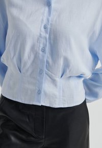 Světlomodrá zkrácená košile na knoflíky s texturovaným povrchem, s řadou knoflíků a jemnými záhyby v oblasti pasu.