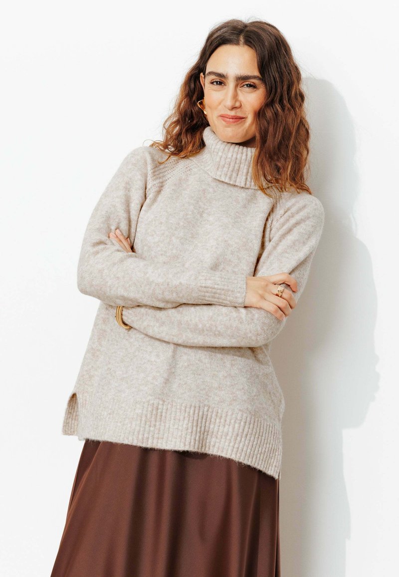 Pull en tricot beige à col roulé avec ourlet côtelé et fentes latérales, porté sur une jupe marron. Texture douce avec une coupe décontractée.
