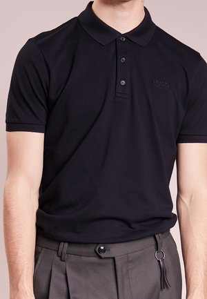 Mann trägt ein eng anliegendes schwarzes Hugo Boss Poloshirt, gesteckt in graue plissierte Hose mit Schlüsselanhänger.