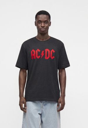 Jeune homme portant un t-shirt noir AC/DC avec un texte rouge et un short en jean bleu clair, debout devant un fond blanc uni.