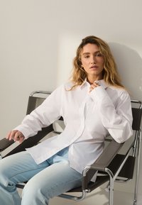 White button-up shirt with long sleeves, relaxed fit. Light blue jeans, seated in a chrome chair with brown cushioned arms. Neutral background.