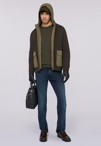 Olivově zelená bunda s kapucí a ribbovanou texturou, se dvěma předními kapsami, nošena přes odpovídající svetr. Modré džíny a černé rukavice dotvářejí vzhled.