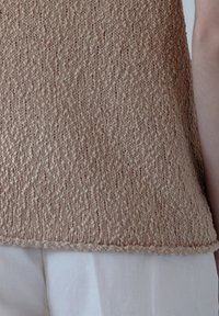 Beżowy sweter z dzianiny o teksturowanej powierzchni i nierównym wzorze ściegów, z prostym dołem i krótkimi rękawami, noszony na białych spodniach.
