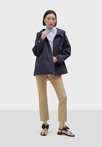 Navy zip-up jacket with a hood, beige fitted pants, and black sandals with bow accents; model stands in a neutral background.