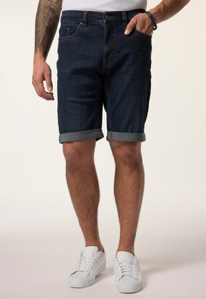 Mann trägt dunkelblaue hochgekrempelte Denim-Shorts, weiße Sneaker und ein weißes Hemd, zeigt tätowierten Unterarm und ein Armband.
