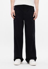 Pantaloni negri cu o croială confortabilă, având pliuri frontale și buzunare, fabricați dintr-un material neted. Asortați cu adidași albi.