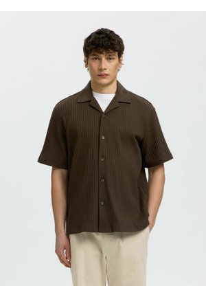 Jeune homme portant une chemise marron côtelée à manches courtes boutonnée sur un débardeur blanc et un pantalon beige, debout devant un fond clair uni.