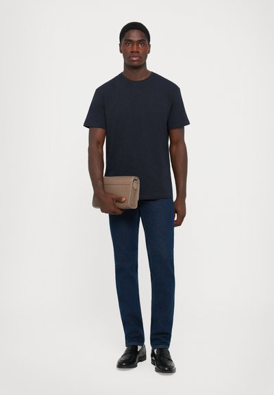 Man standing wearing a dark navy t-shirt, blue jeans, black shoes, holding a taupe leather clutch bag against a plain background.