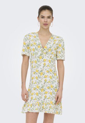 Giovane donna che indossa un vestito bianco con motivo di limoni gialli e foglie verdi, maniche corte, scollatura a V e orlo arricciato, in piedi.