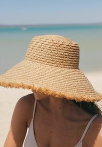 Sombrero de paja de ala ancha con un diseño tejido texturizado y borde deshilachado, en tonos naturales, que parcialmente oculta la cara contra un fondo de playa.