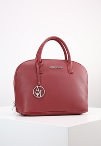 Červená syntetická kabelka se strukturovaným designem, dvojitými uchy, stříbrnými kovovými detaily, embosovaným logem a odnímatelným přívěskem s logem.