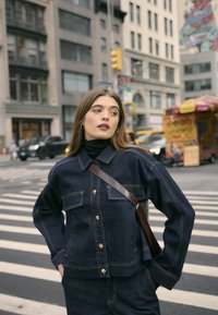 Tumedat denimpusaak kuldsete nappidega, kahte rinnataskut ja kontrastset kollast õmblust, kombineeritud musta kõrge kaelusega särgiga.