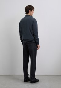 Joven de pie de lado, vistiendo un suéter gris oscuro, pantalones negros y zapatos negros, contra un fondo gris claro liso.