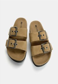 Bruine suède instapsandalen met twee brede bandjes en verstelbare metalen gespen, een textuur van de voetbed, en een zwarte rubberen zool.