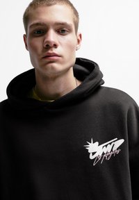 Črna kapuca s sprednjo nalepko, na kateri je napis "Los Angeles" v belem in roza pisavi. Material izgleda mehak; dizajn vključuje velik kapuco.