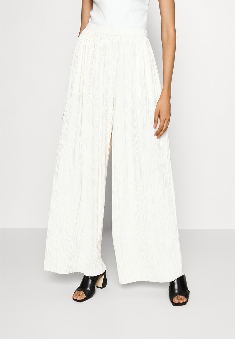 Plisserade, högmidjade vida byxor i off-white tyg, med en frontslits och en mjuk, texturerad yta. Matchas med svarta öppen-tå skor.