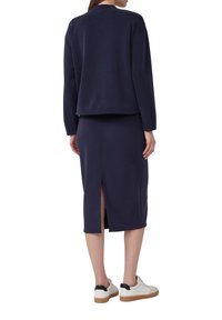 Navyblauer, figurbetonter Pullover und Midirock-Set, mit einer Rückenschlitzdetail. Das Material wirkt glatt mit einer weichen Textur. Weiße Sneakers runden den Look ab.