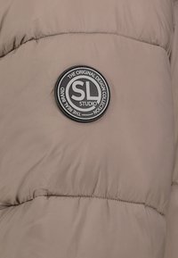 Pufferjack in lichtbruin stof met een gladde textuur. Heeft een ronde zwarte logo patch met witte tekst aan de voorkant.