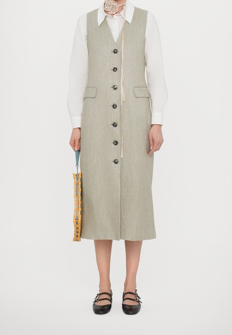 Femme portant une robe beige sans manches boutonnée sur une chemise blanche, tenant un petit sac à main jaune et bleu, et portant des chaussures noires à double bride.